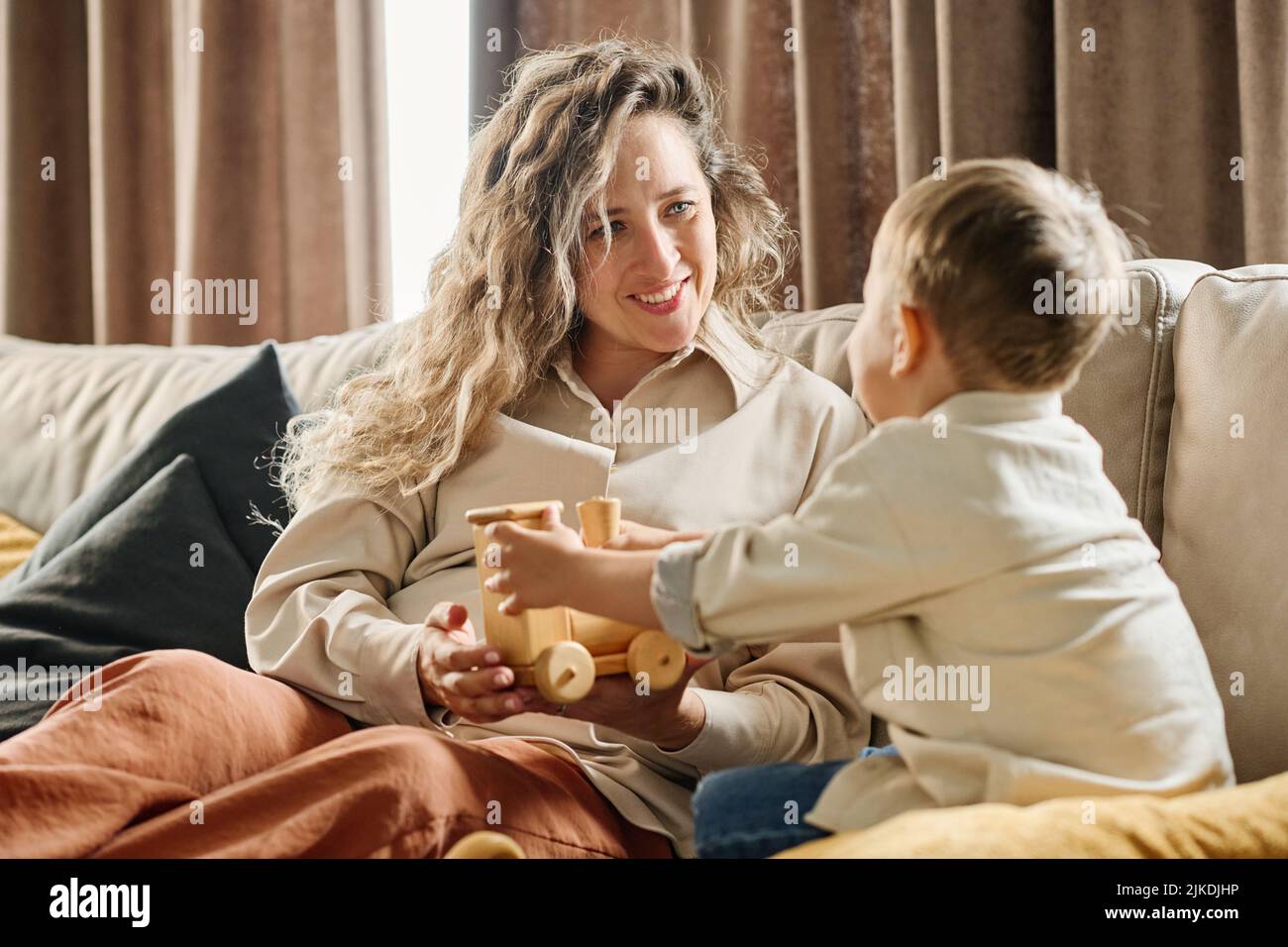 Felice madre giovane guardando il suo piccolo figlio con sorriso mentre lo mostra nuovo giocattolo di legno durante il riposo sul divano in salotto Foto Stock