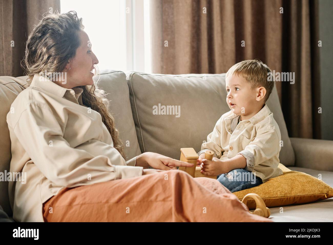 Giovane donna incinta seduta sul divano di fronte al figlio piccolo che tiene un nuovo giocattolo di legno e parla con lui mentre passa il tempo insieme Foto Stock