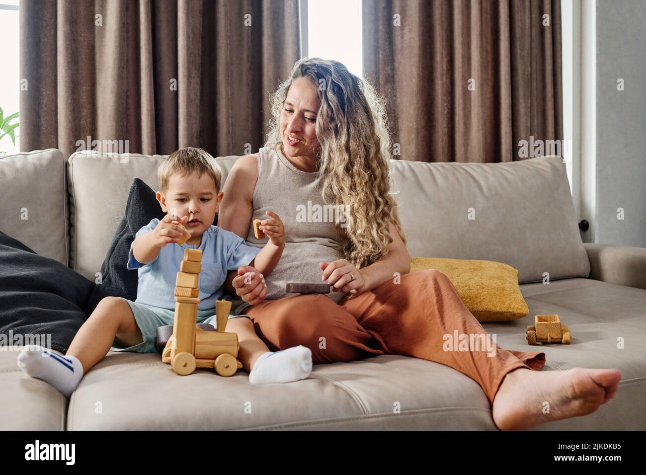 Carino ragazzino che mette la pila di cubi di legno sulla parte superiore della locomotiva giocattolo mentre si siede accanto a sua madre su un ampio comodo divano Foto Stock