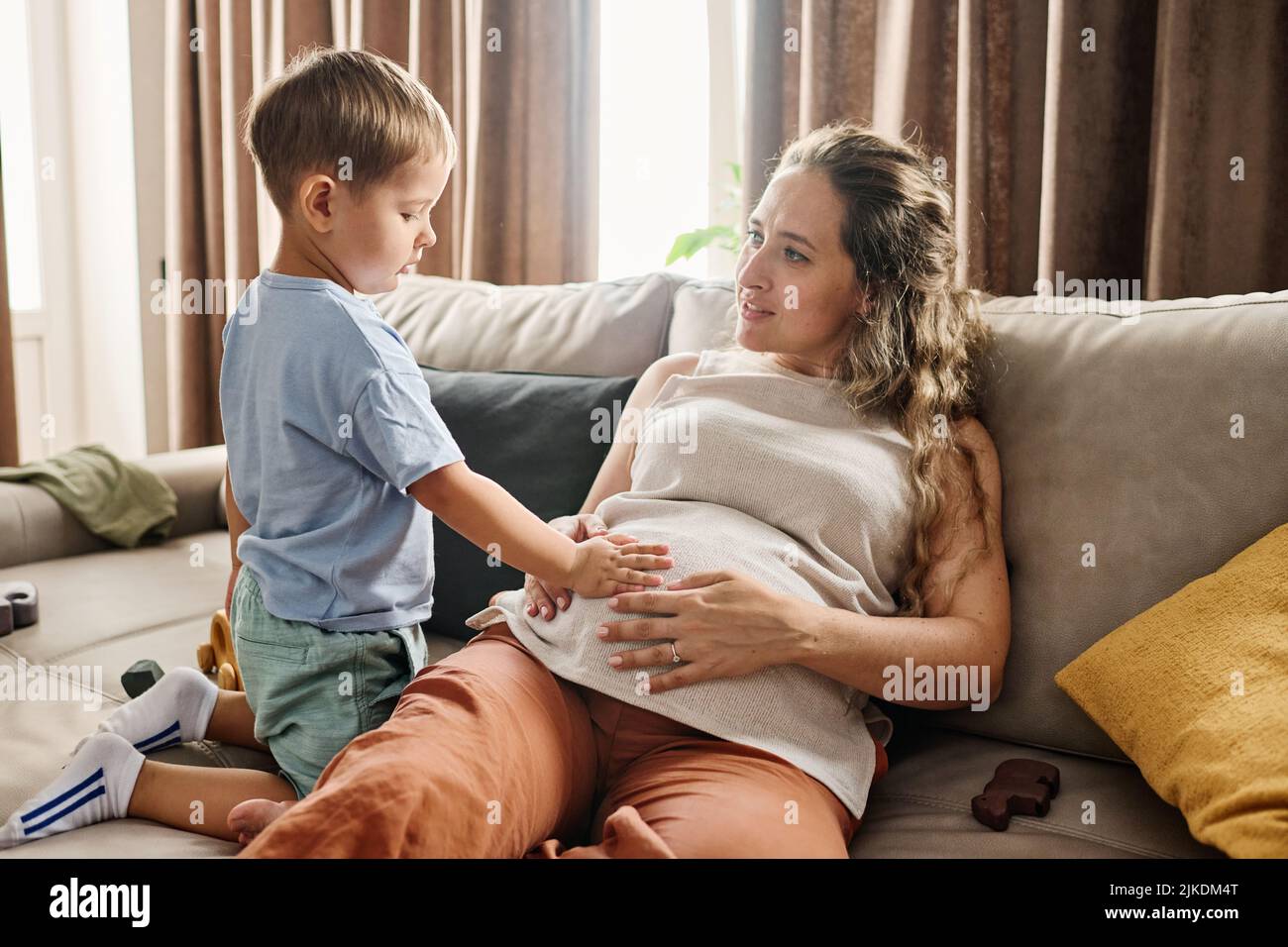 Curioso toddler tenere la mano sul ventre della sua madre incinta seduta sul divano mentre spende il tempo libero con il suo piccolo figlio carino a casa Foto Stock
