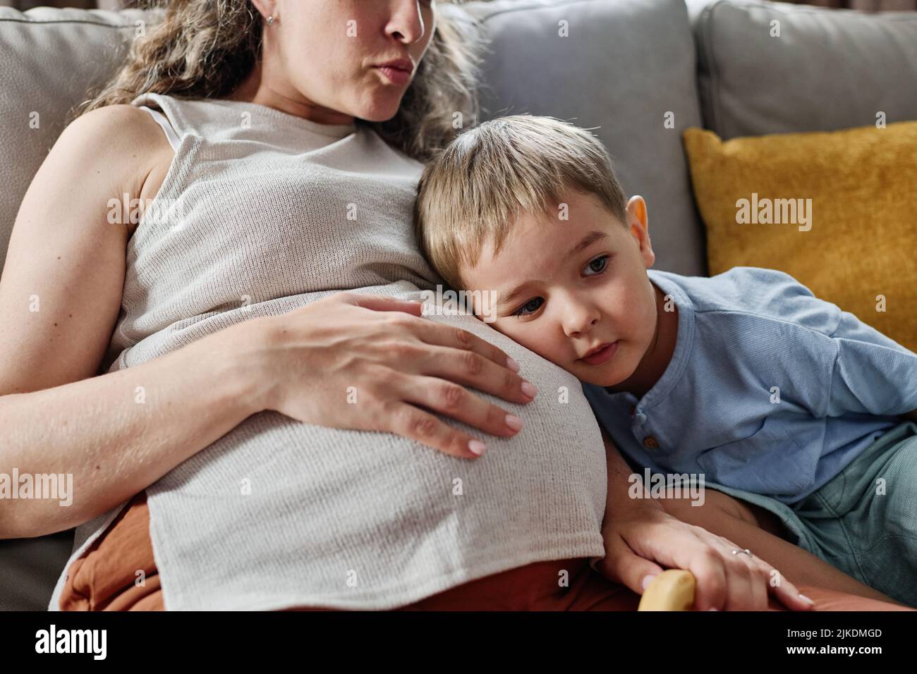 Curioso ragazzino che tiene la testa sul ventre della madre incinta mentre ascolta il battito cardiaco del feto in grembo mentre si siede sul divano Foto Stock