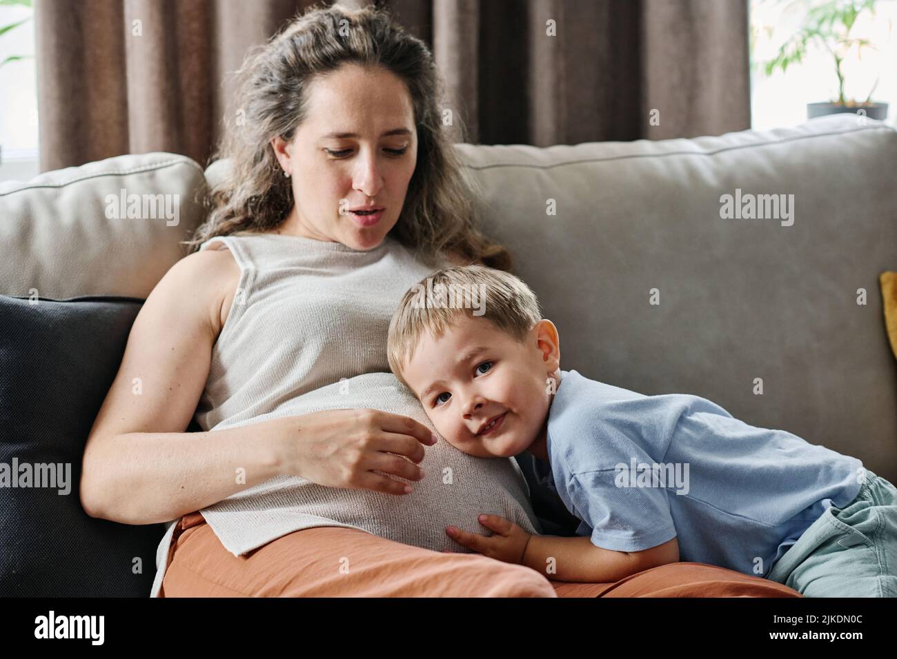 Carino ragazzo in casualwear tenendo l'orecchio vicino al ventre della sua madre incinta mentre cerca di catturare il battito cardiaco del futuro bambino Foto Stock