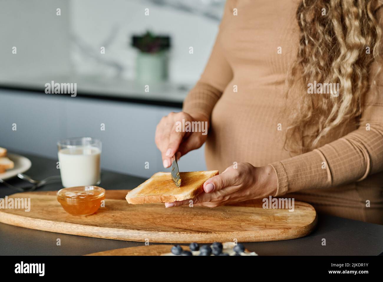 Mani di giovane donna incinta con coltello che sparge miele o marmellata su fetta di pane fresco di grano mentre si prepara la colazione Foto Stock