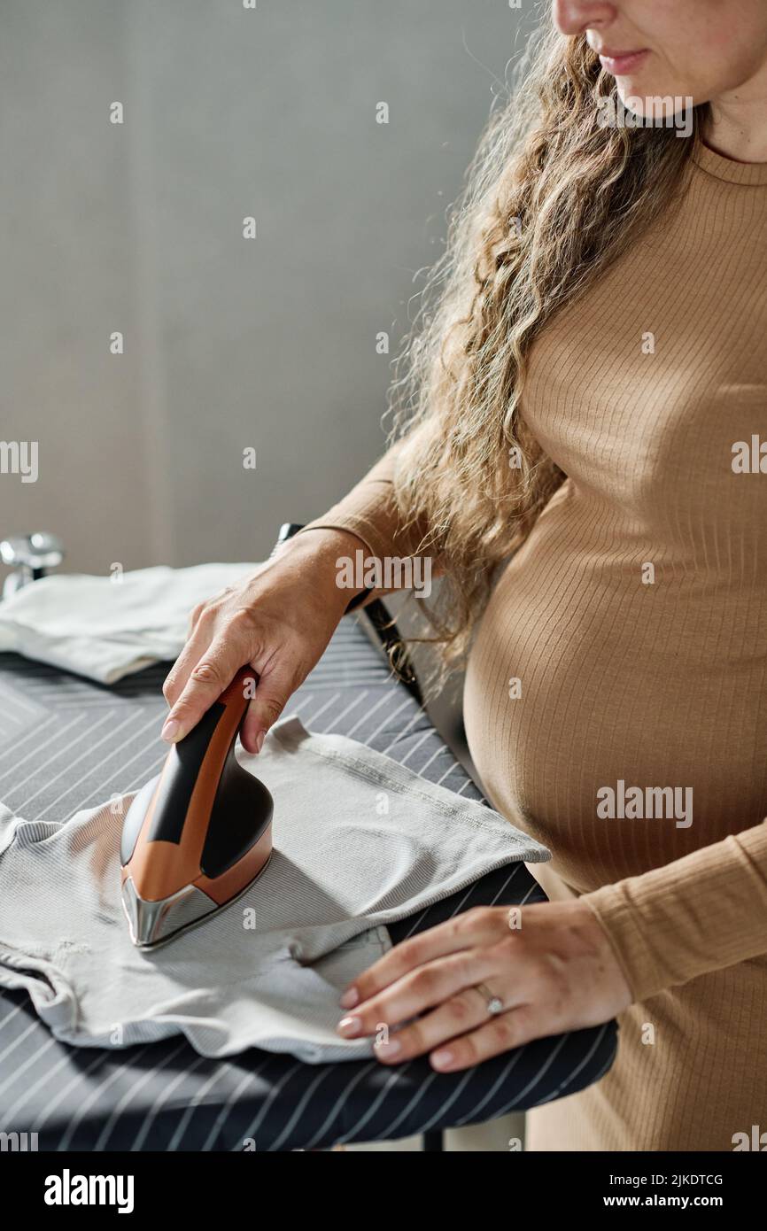 Giovane donna incinta in abito casual di vestiti da stiro di colore beige del suo bambino con ferro elettrico mentre si trova a bordo Foto Stock