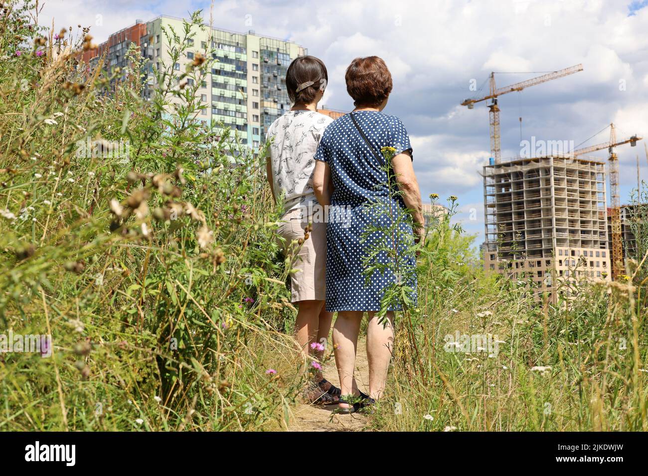Due donne in piedi sul prato verde e guardando le nuove case del quartiere residenziale e gru edili. I compratori scelgono l'appartamento Foto Stock