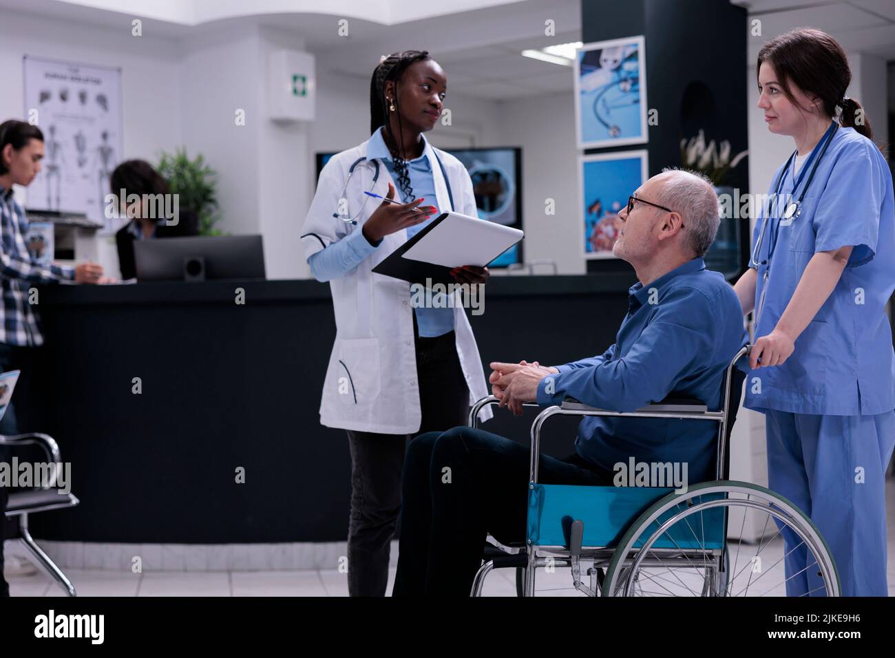Medico afroamericano che tiene appunti con i dati medici che parlano con l'infermiera circa il paziente anziano in sedia a rotelle nella ricezione dell'ospedale. Uomo anziano che vive con disabilità in clinica per appuntamento. Foto Stock