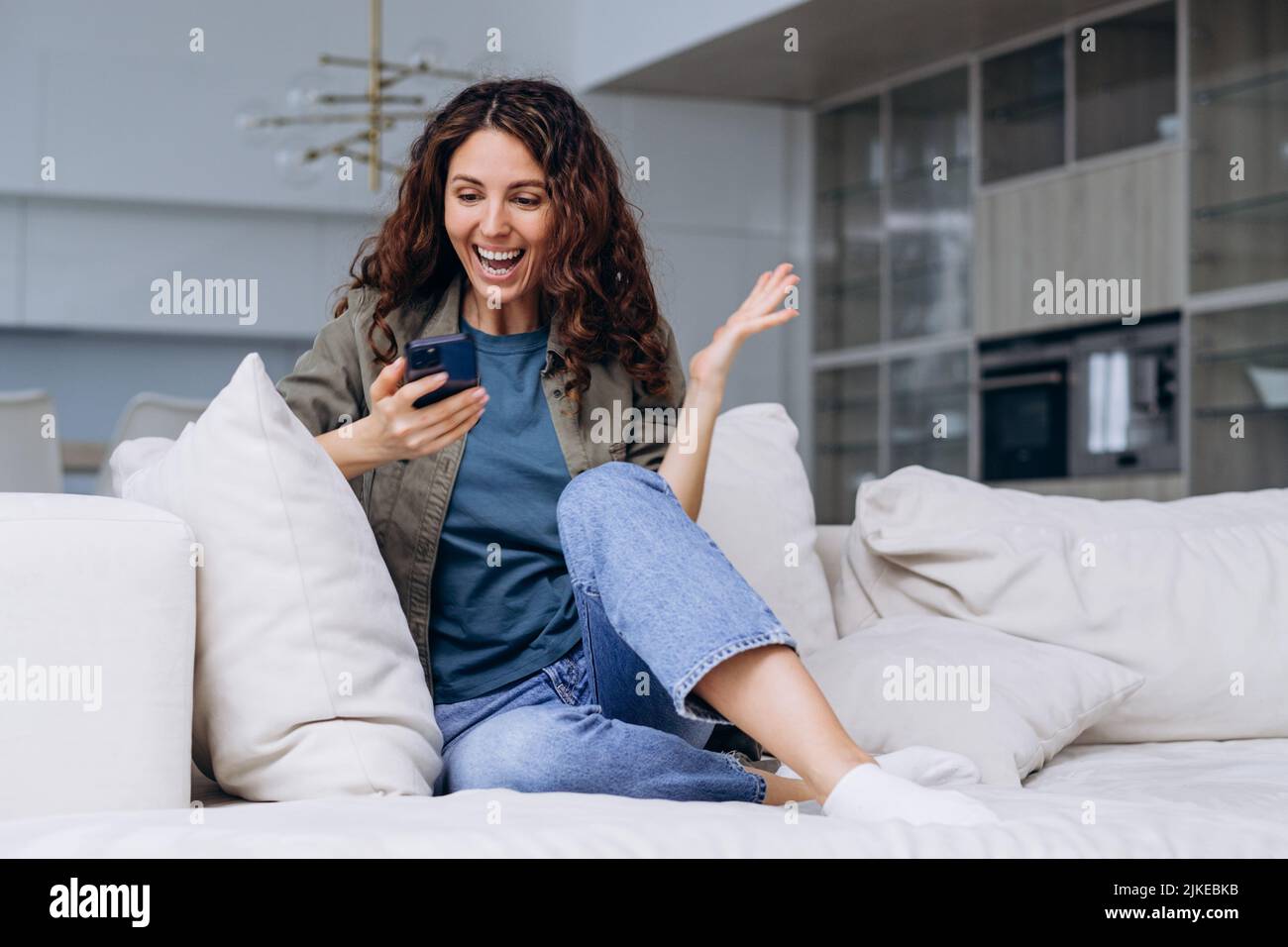 Gioiosa donna ricciola con un telefono nelle sue mani ha sollevato la sua mano da una notizia molto piacevole a casa seduta sul divano, in attesa di una promozione sul lavoro o ha ricevuto un grande ordine Foto Stock