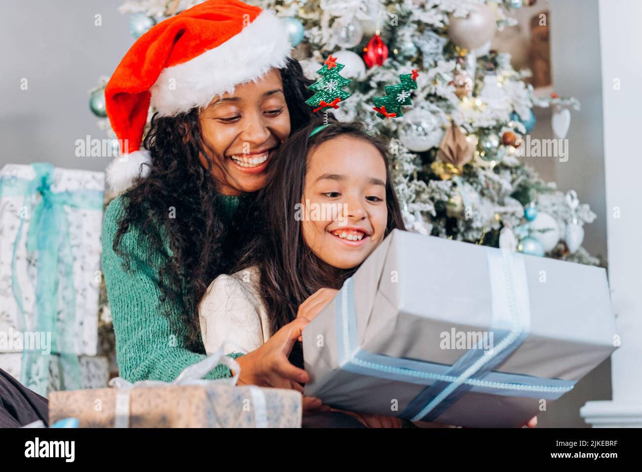 Curly African American donna e bruna ragazza dai capelli lunghi che indossa i cappelli di festa aprire le scatole presenti con sorriso che siede all'albero di Natale Foto Stock