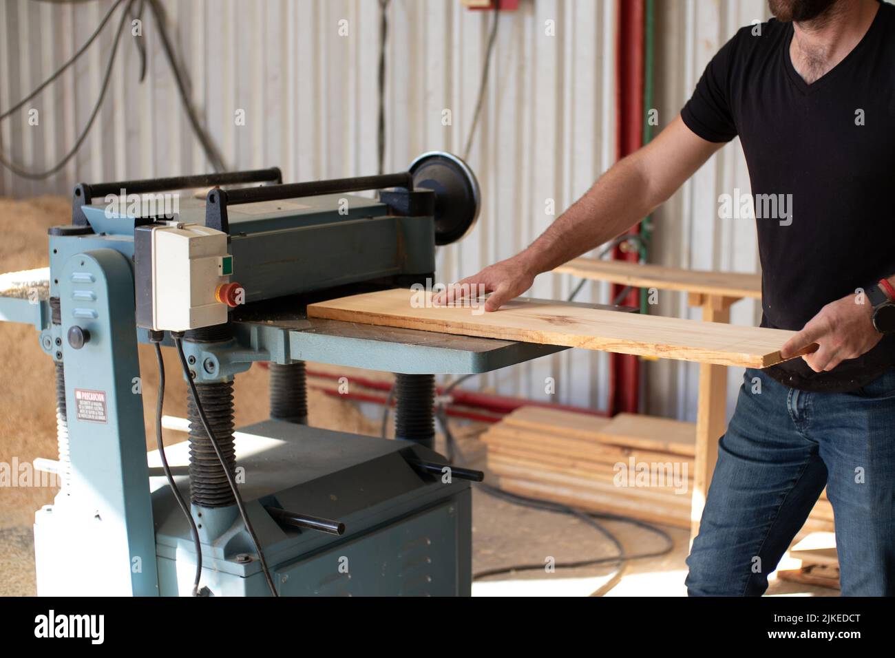 Giovane carpentiere che lavora su una macchina elettrica da banco Jointer che taglia un asse di legno in un'officina di carpenteria usando guanti neri di sicurezza Foto Stock