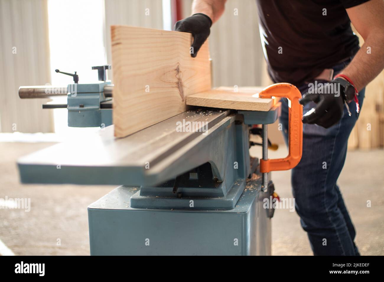 Giovane carpentiere che lavora su una macchina elettrica da banco Jointer che taglia un asse di legno in un'officina di carpenteria usando guanti neri di sicurezza Foto Stock