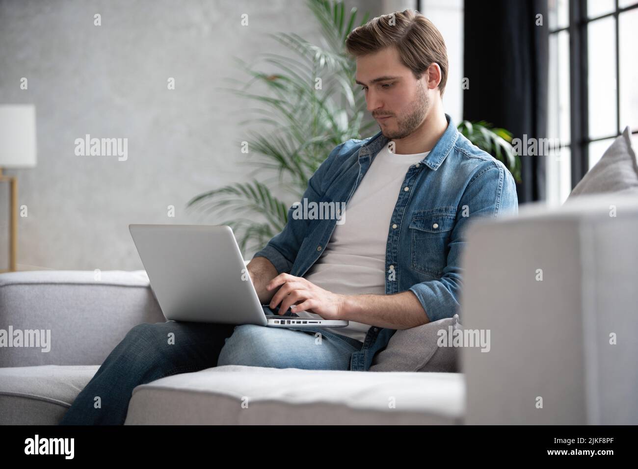 Giovane uomo sorridente e attraente sta navigando al suo laptop, seduto a casa sul comodo divano a casa, indossando abiti casual Foto Stock