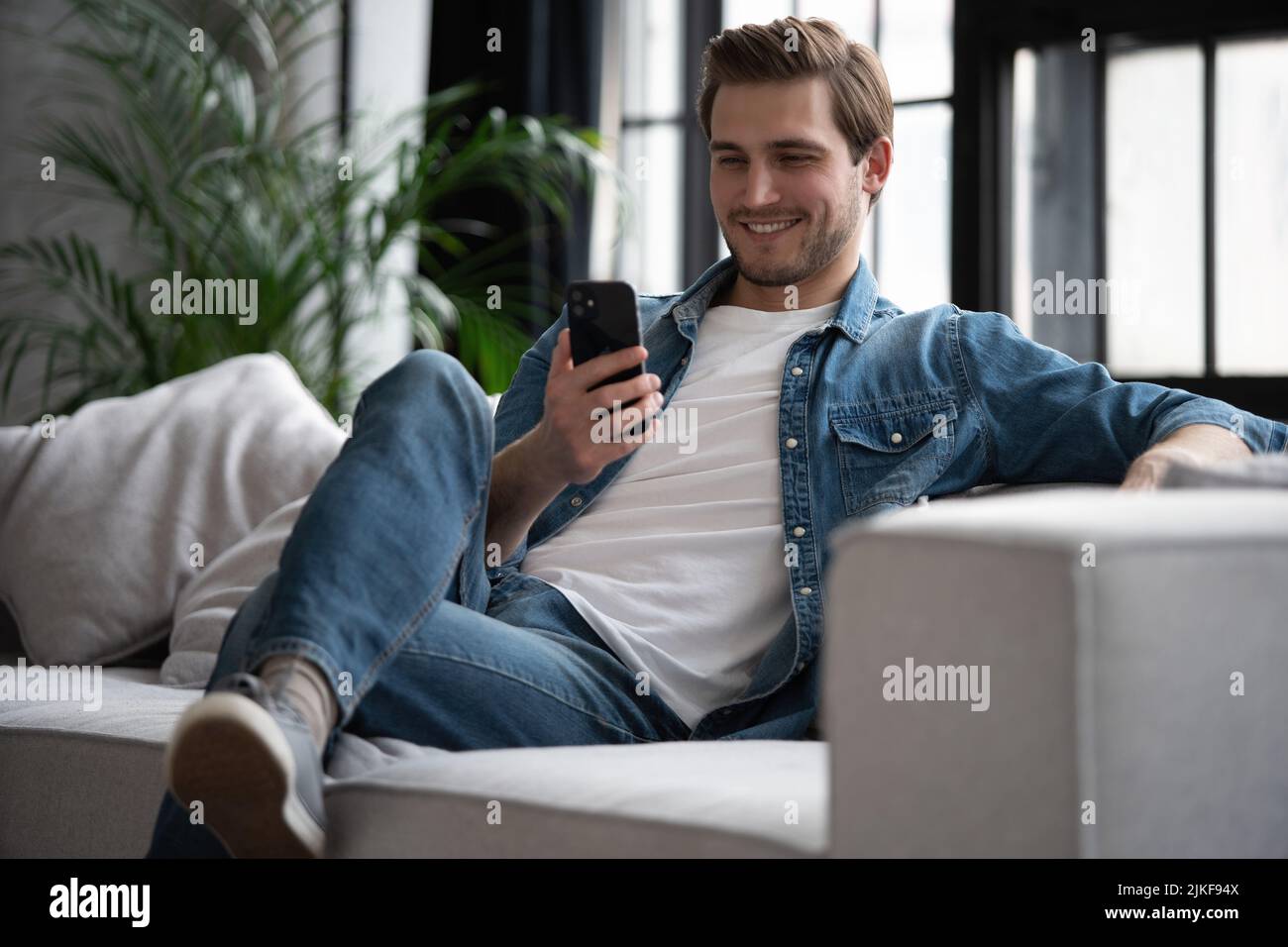 Un giovane attraente e sorridente che indossa abiti casual seduti su un divano nel soggiorno, utilizzando il telefono cellulare Foto Stock
