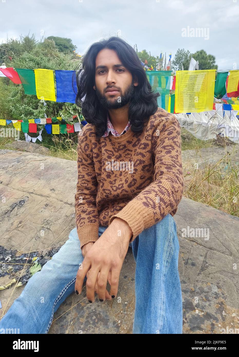 Un giovane uomo con capelli lunghi e barba che guarda la macchina fotografica mentre si siede sulla roccia con lo sfondo delle bandiere tibetane di preghiera a Rewalsar Foto Stock