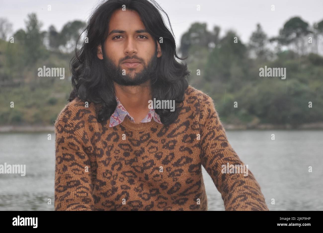 Primo piano di un giovane ragazzo indiano attraente con capelli lunghi e barba guardando la macchina fotografica mentre si siede accanto al lago Foto Stock