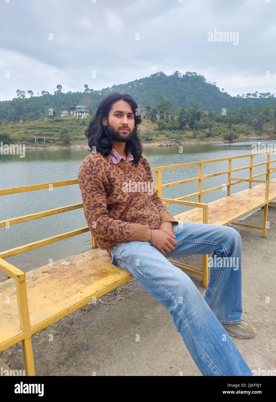 Un giovane indiano dall'aspetto buono con capelli lunghi e barba che guarda la macchina fotografica mentre si siede sulla panca accanto al lago Foto Stock