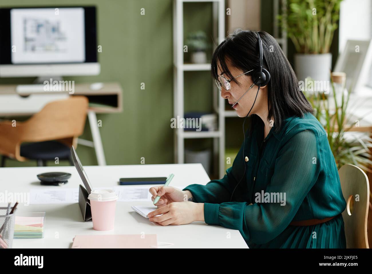 Giovane donna d'affari asiatica in cuffia per prendere appunti seduti sul posto di lavoro con il tablet pc durante la conversazione online Foto Stock