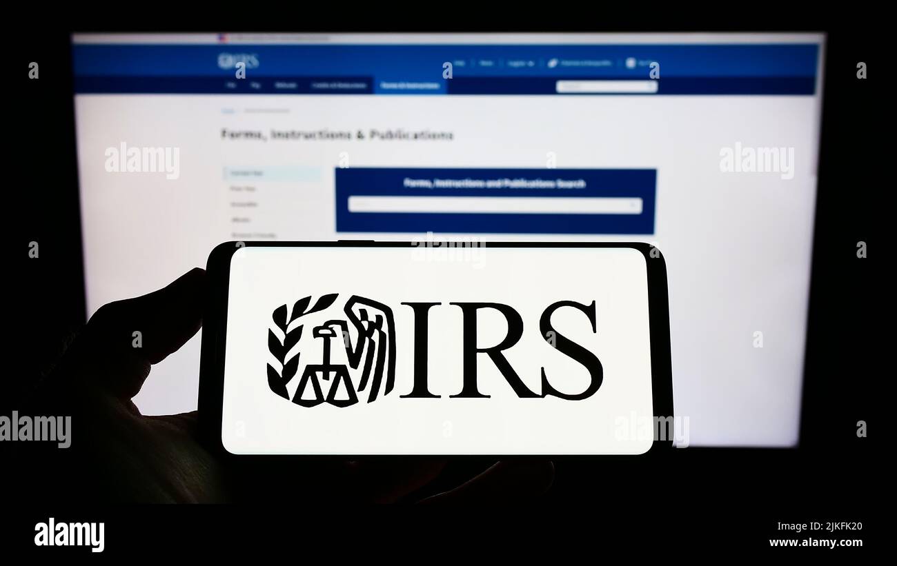 Persona che tiene uno smartphone con il logo dell'IRS (US Internal Revenue Service) sullo schermo di fronte al sito Web. Mettere a fuoco sul display del telefono. Foto Stock