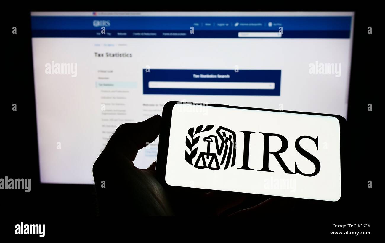 Persona che tiene il cellulare con il logo di US Internal Revenue Service (IRS) sullo schermo di fronte alla pagina web. Mettere a fuoco sul display del telefono. Foto Stock