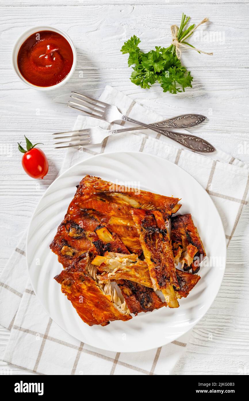 Cadere fuori le costole di forno di osso cotti su piastra bianca con ketchup su tavola di legno bianco, vista verticale dall'alto Foto Stock