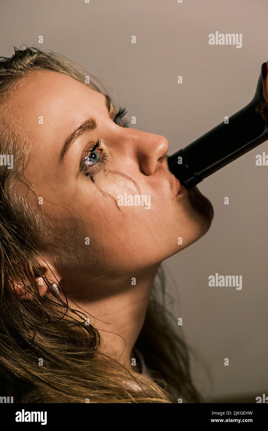 Piangendo la giovane donna con il mascara macchiato beve alcol dal collo di bottiglia. Vista laterale ritratto di donna infelice che sperimenta crisi emotiva nella vita Foto Stock