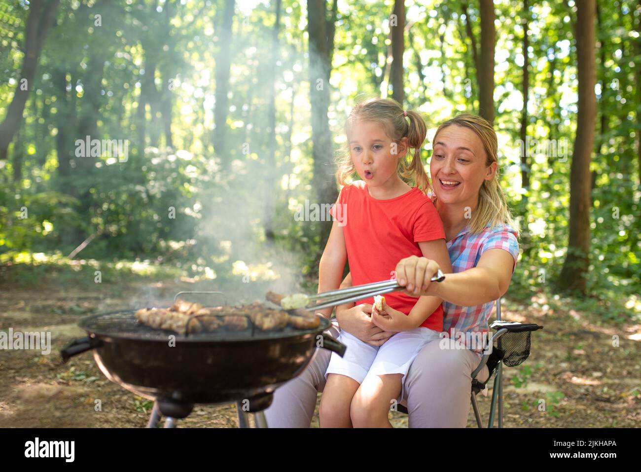 Una giovane madre felice campeggio e cucina barbecue in una foresta con sua figlia Foto Stock