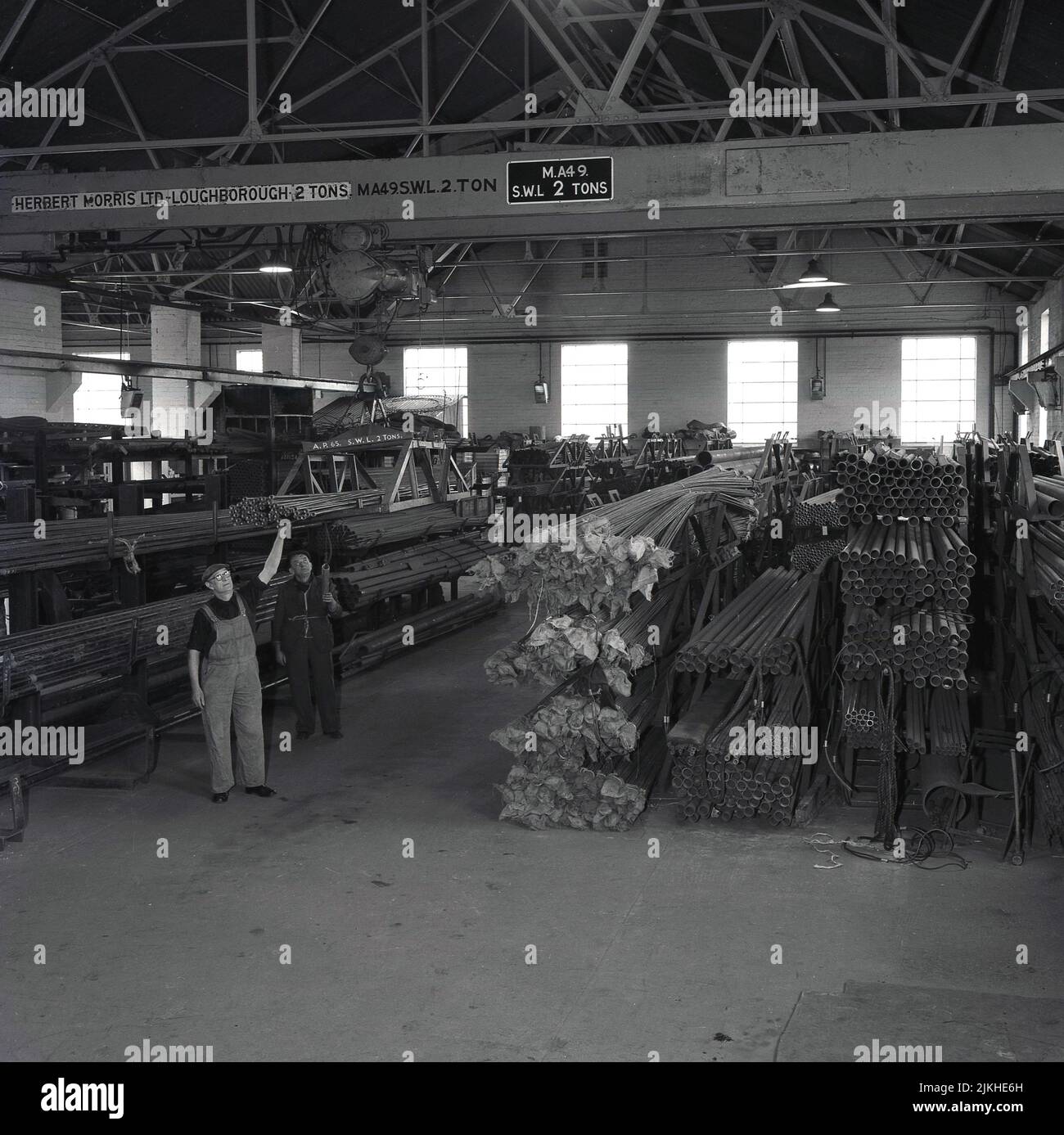 1950s, storico, mucchi di aste d'acciaio in un grande capannone o appendiabiti, Port Talbot, Galles, Regno Unito. Due operai siderurgici che utilizzano macchinari per il sollevamento di carroponte prodotti da Herbert Morris Ltd di Loughborough - produttori specializzati di gru e blocchi-puleggia - per sollevare e manovrare le tubazioni. Niente cappelli duri, solo cappelli di stoffa! Foto Stock