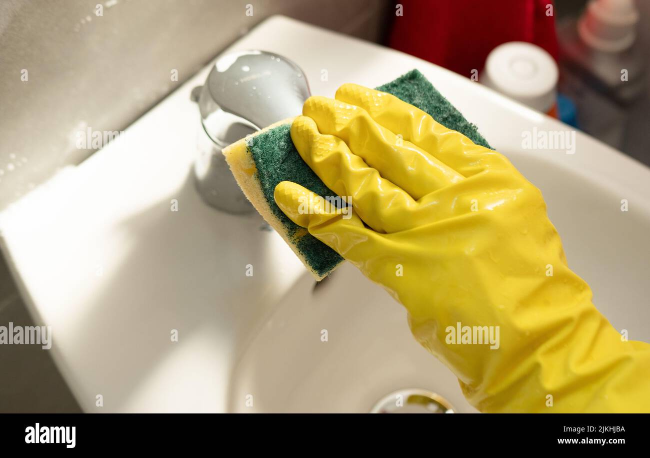 Ripulire la casa. Donna che fa i chores in bagno, mani in guanti gialli pulizia del rubinetto d'acqua, lavello in acciaio con straccio blu e spray detergente Foto Stock