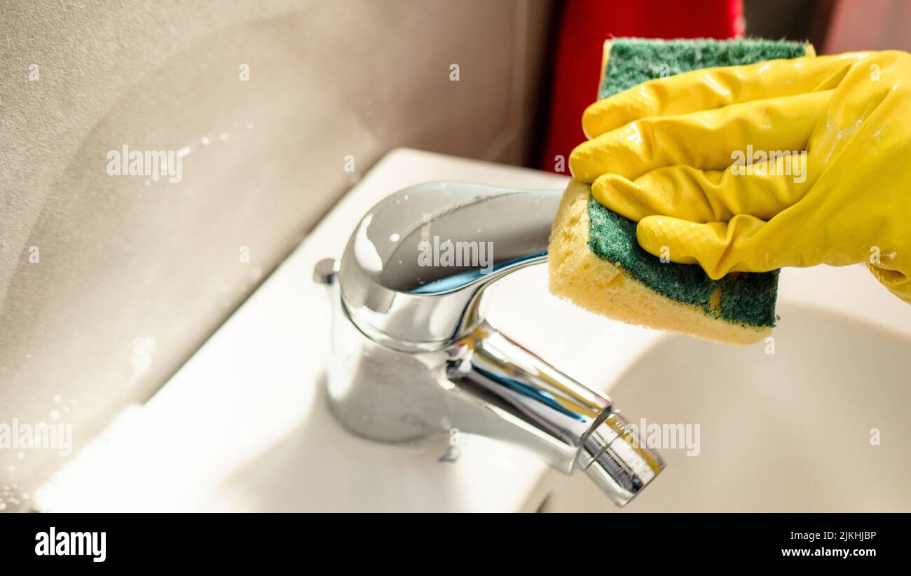 Ripulire la casa. Donna che fa i chores in bagno, mani in guanti gialli pulizia del rubinetto d'acqua, lavello in acciaio con straccio blu e spray detergente Foto Stock