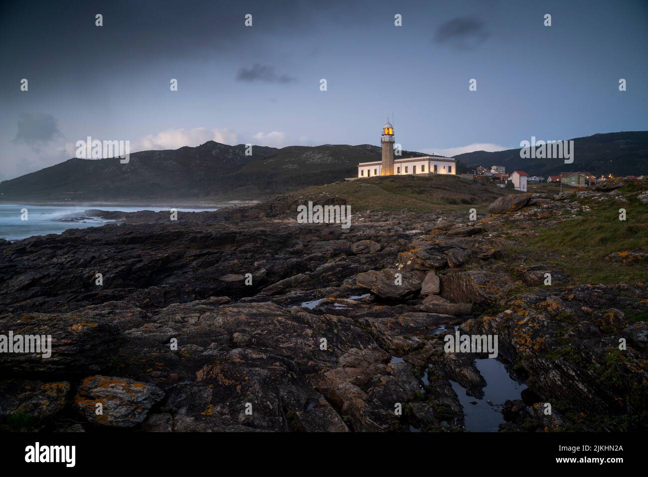 Uno scatto orizzontale di un faro in Galizia dietro un paesaggio roccioso sotto un cielo cupo Foto Stock