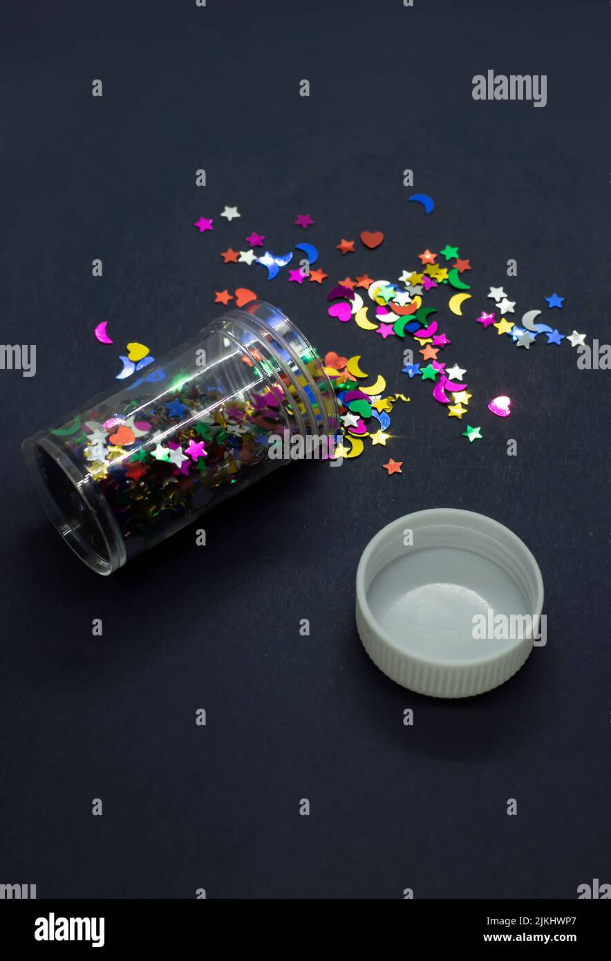 Un primo piano di un contenitore pieno di glitter scintilla in forme diverse sparso sulla superficie grigia Foto Stock