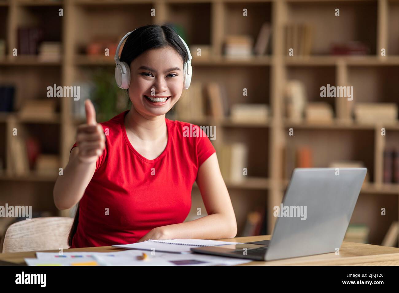 Felice giovane donna asiatica con sindrome di Down in cuffie con dito puntino portatile alla fotocamera in ufficio Foto Stock