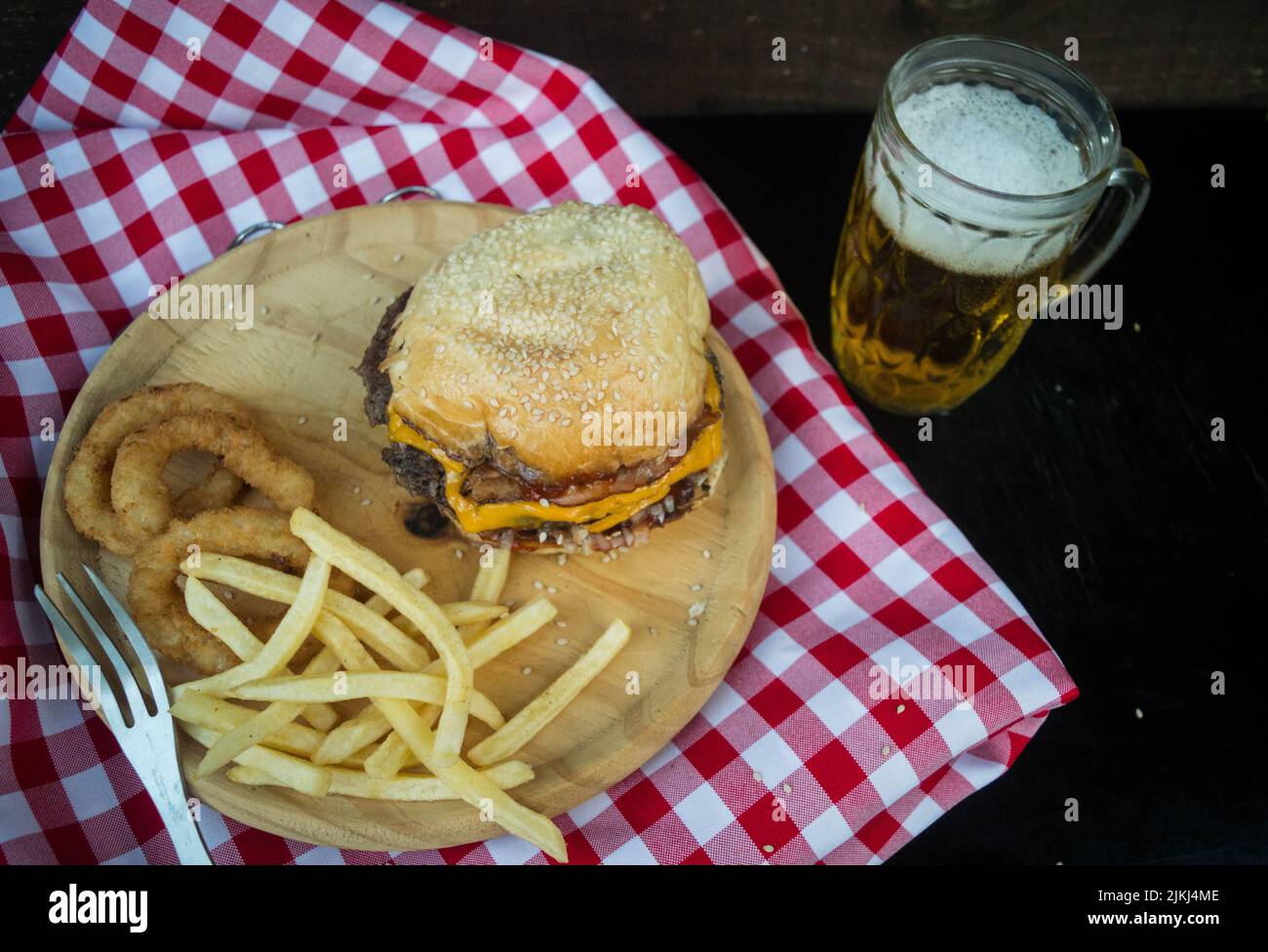 Un angolo di un hamburger su un piatto di legno con anelli di cipolla e patatine fritte accanto a un bicchiere di birra. Foto Stock