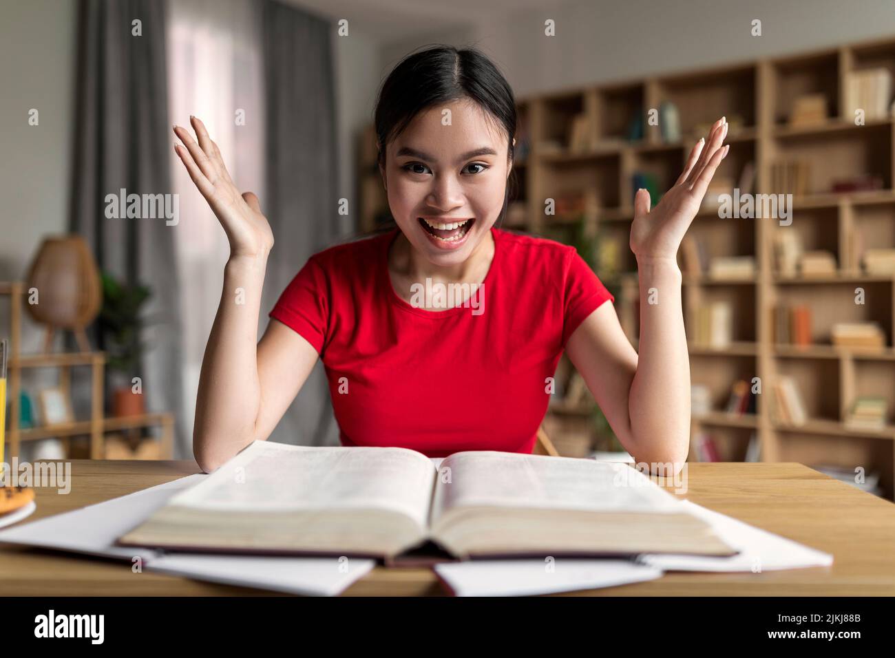 Studio a casa due covid-19. Soddisfatto eccitato giovane studentessa asiatica con libro urlando e gioisce Foto Stock