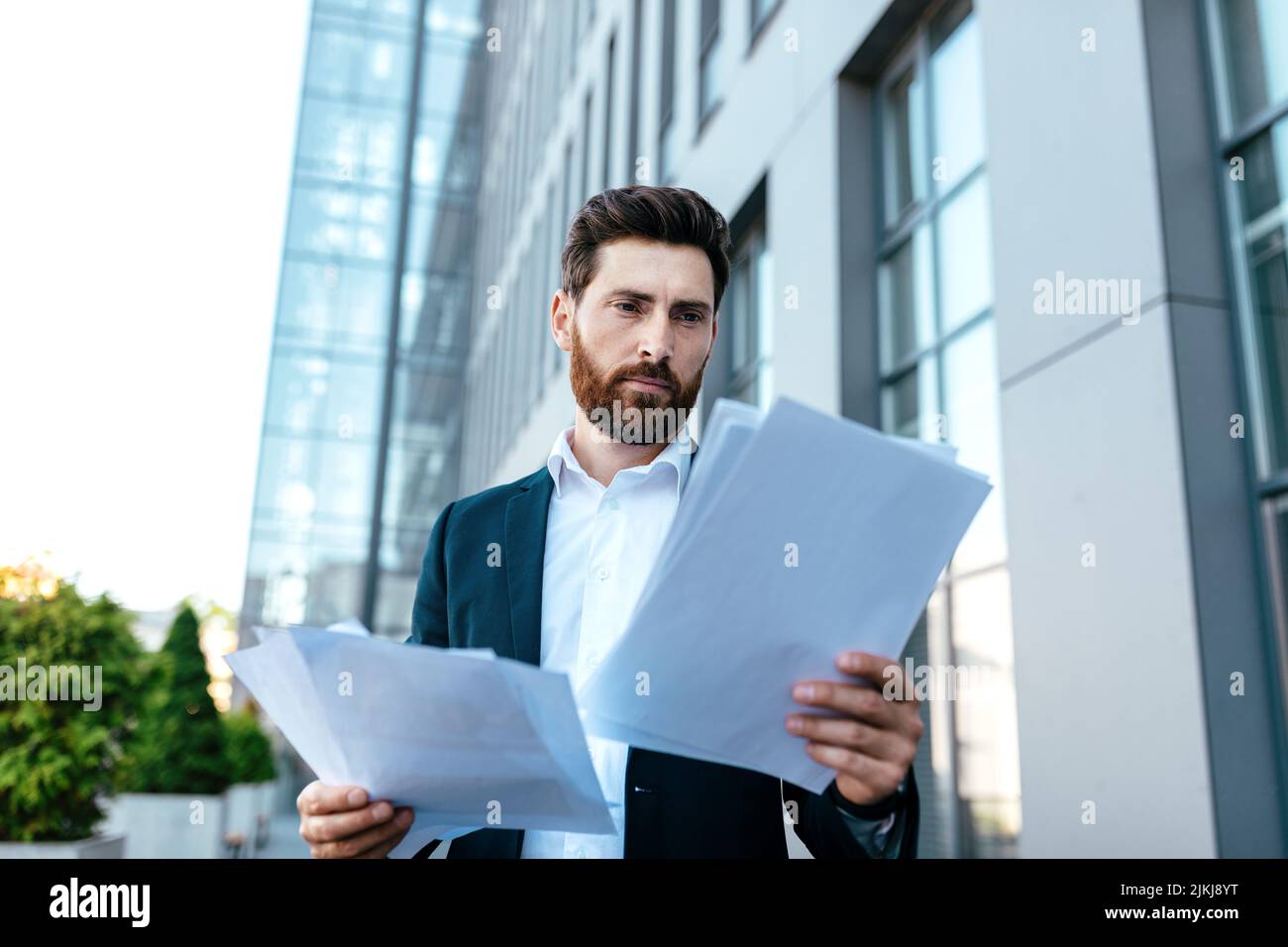 Occupato uomo d'affari giovane sicuro con la barba in vestito lavora con documenti vicino ufficio edificio Foto Stock