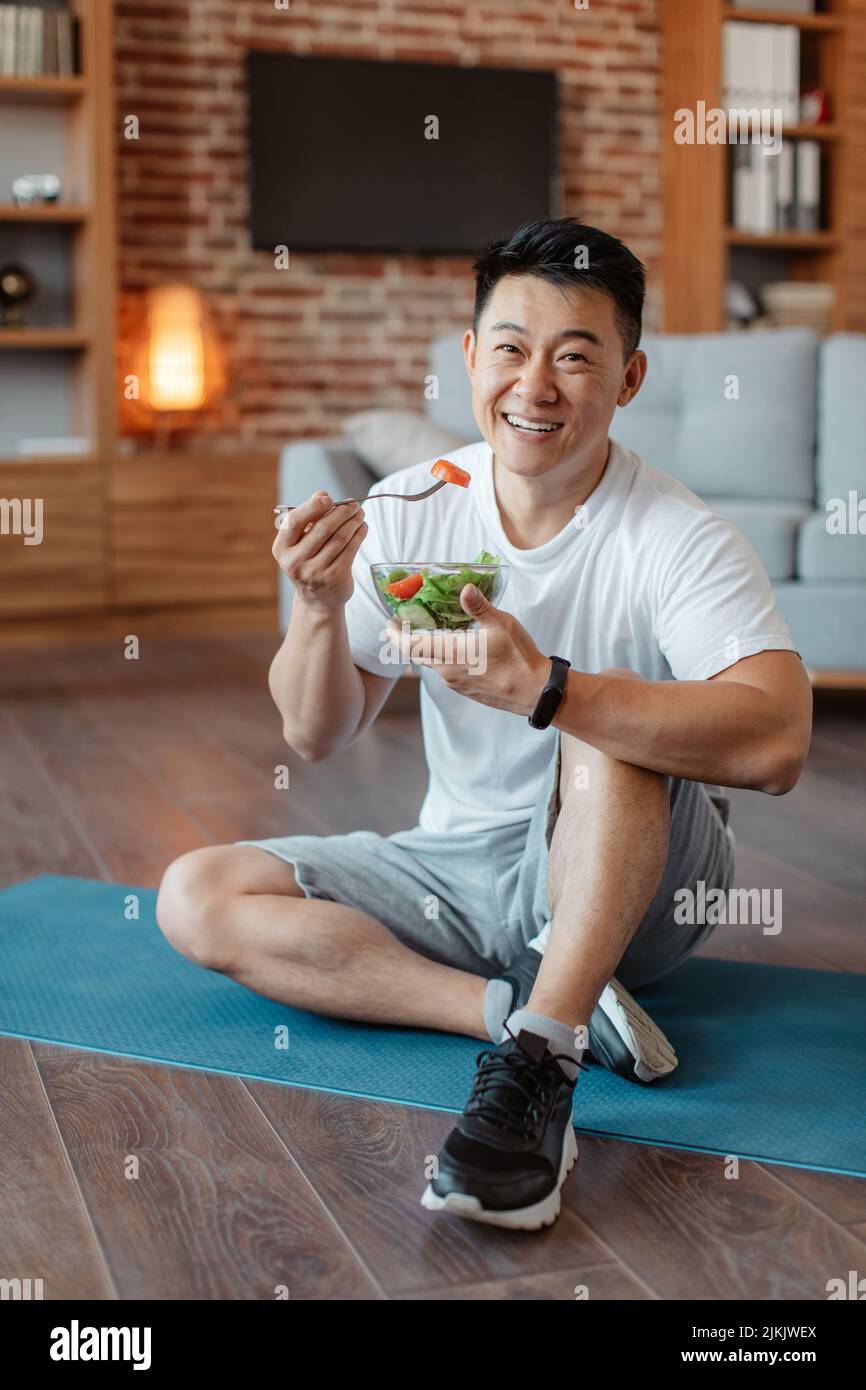 Concetto di stile di vita sano. Felice uomo asiatico di mezza età che mangia insalata di verdure, seduto sul tappeto yoga e sorridente alla macchina fotografica Foto Stock