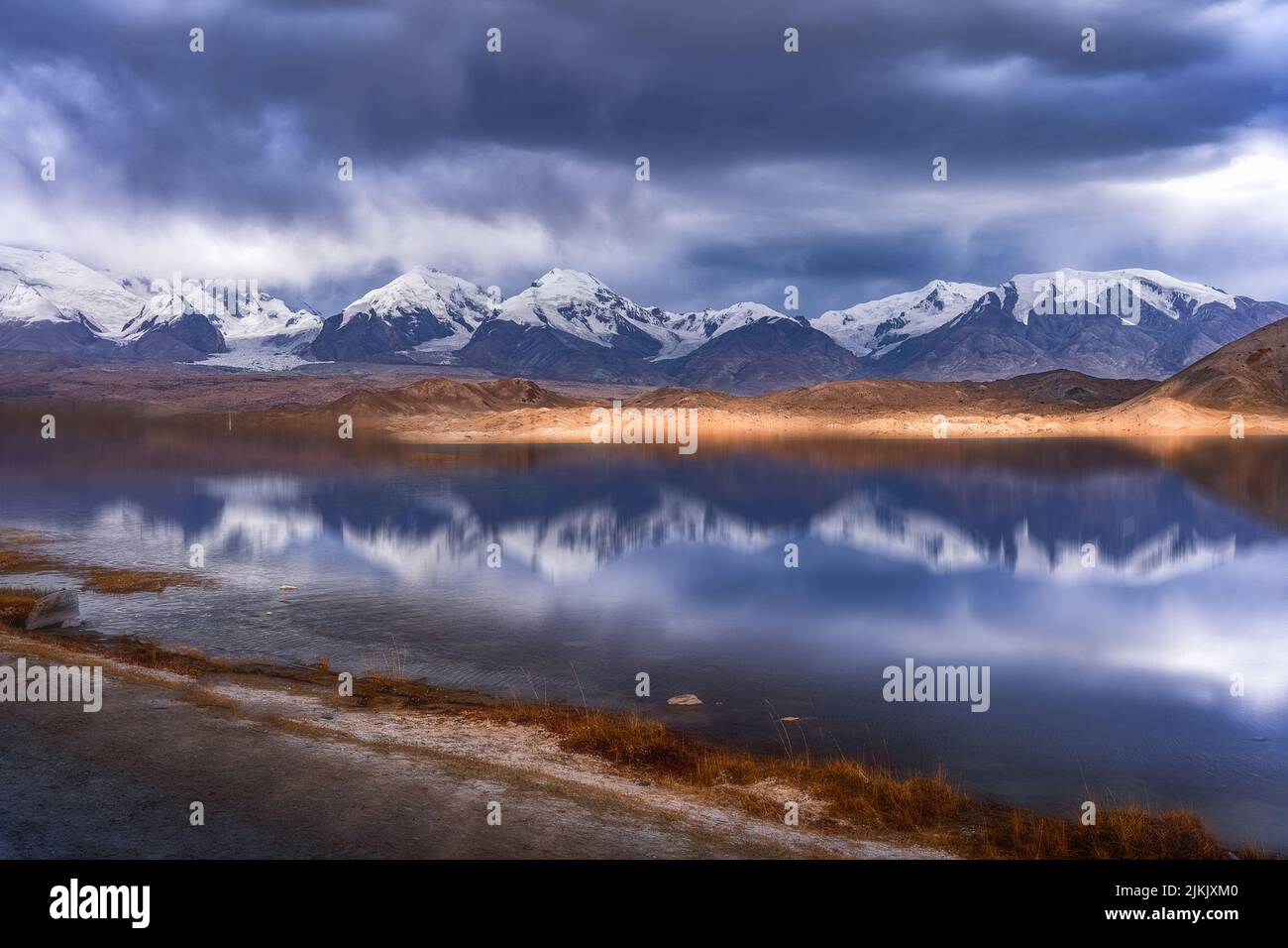 Una vista panoramica della cima del Kongur Tobe Feng sulla riva del lago riflettente sotto il cielo cupo Foto Stock