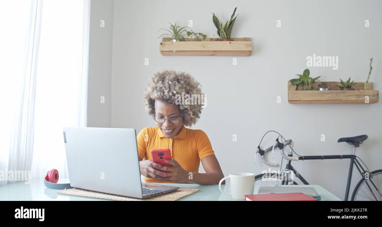 Una donna africana riccia che guarda il suo smartphone in ufficio davanti a un computer portatile Foto Stock