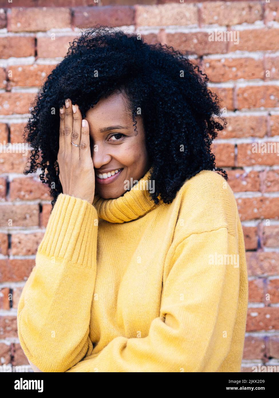 Una donna africana dalla Spagna in maglione giallo che copre la faccia con mano con muro di mattoni sullo sfondo Foto Stock Una donna africana dalla Spagna in maglione giallo che copre la faccia con mano con muro di mattoni sullo sfondo Foto Stock