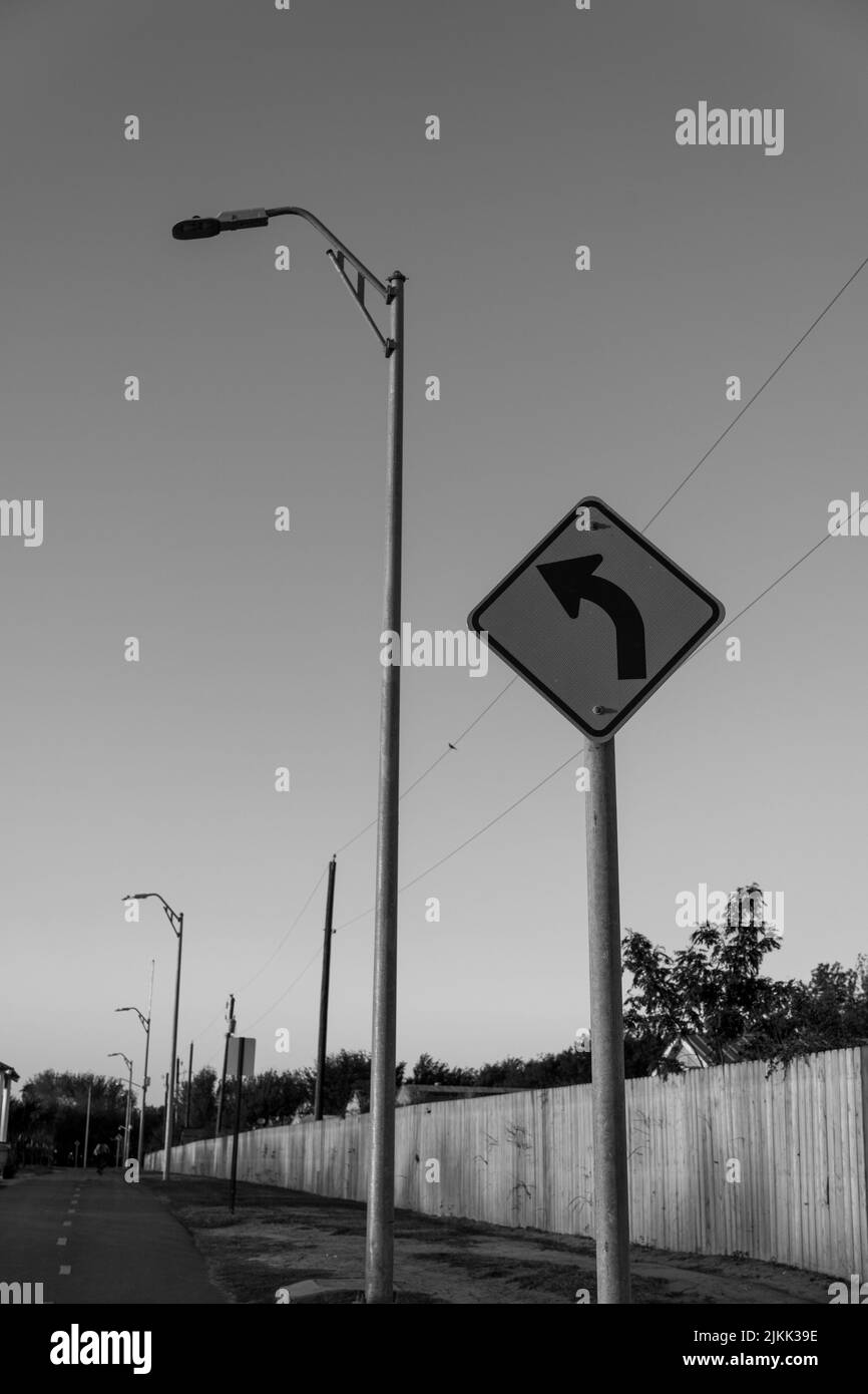 Immagine in scala di grigi di un cartello stradale con curva a sinistra in avanti su un palo della strada accanto a un lampione Foto Stock