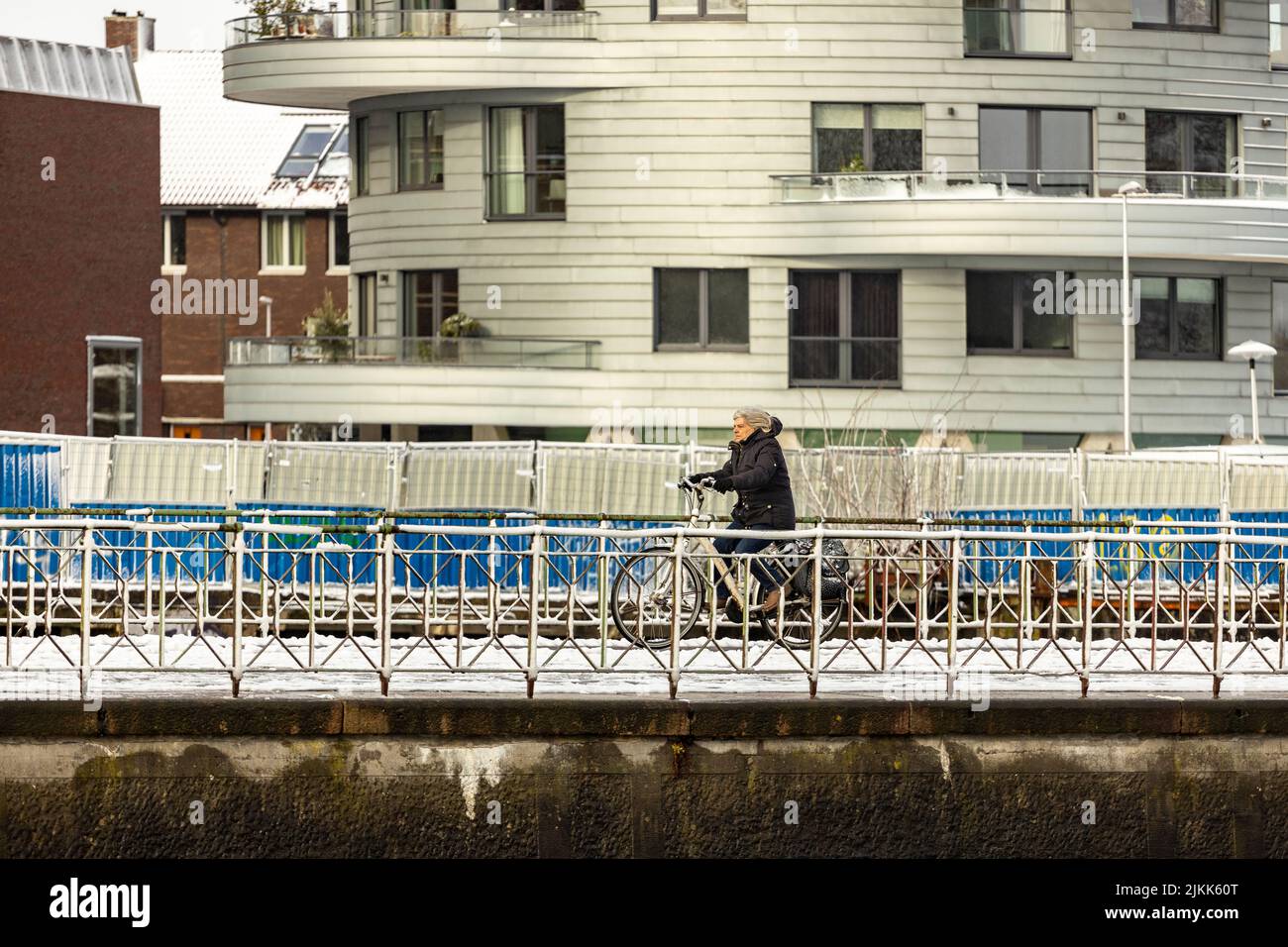 Donna in bicicletta su una banchina innevata con moderna torre residenziale contemporanea sullo sfondo. Condizioni meteorologiche olandesi e trasporto Foto Stock