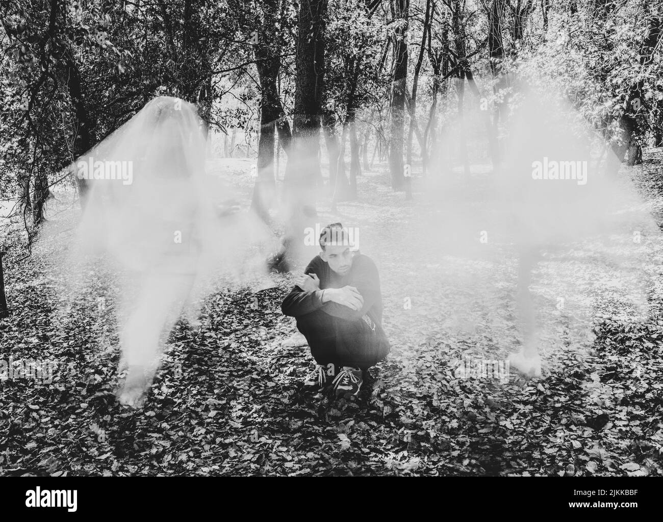 Un colpo in scala di grigi di un uomo che indossa un nero pieno seduto a terra con una nebbia bianca davanti a lui nel mezzo di una foresta Foto Stock