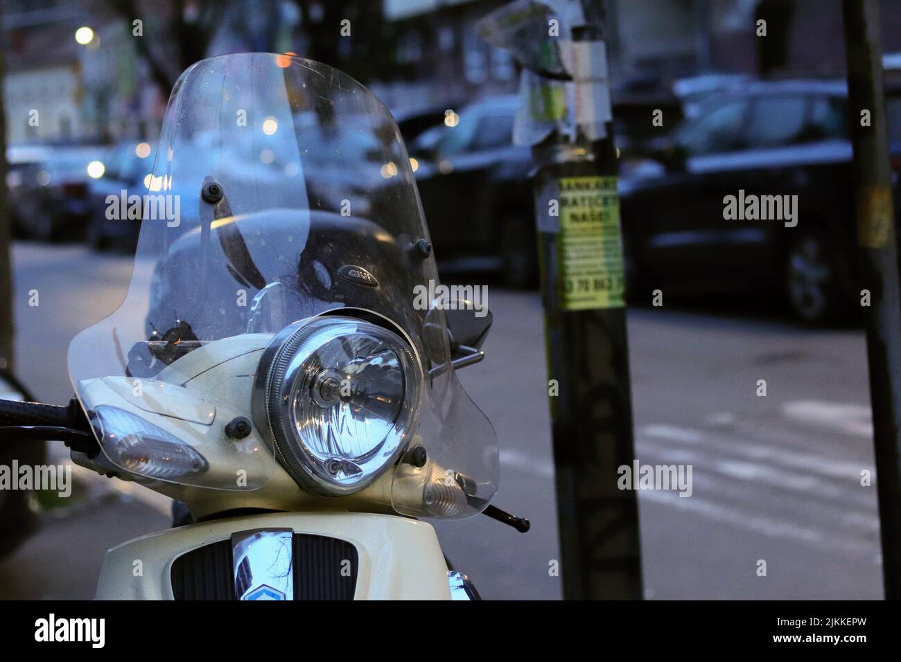 Un primo piano di uno scooter vespa d'epoca parcheggiato in strada al tramonto con una fila di auto parcheggiate sullo sfondo sfocato Foto Stock