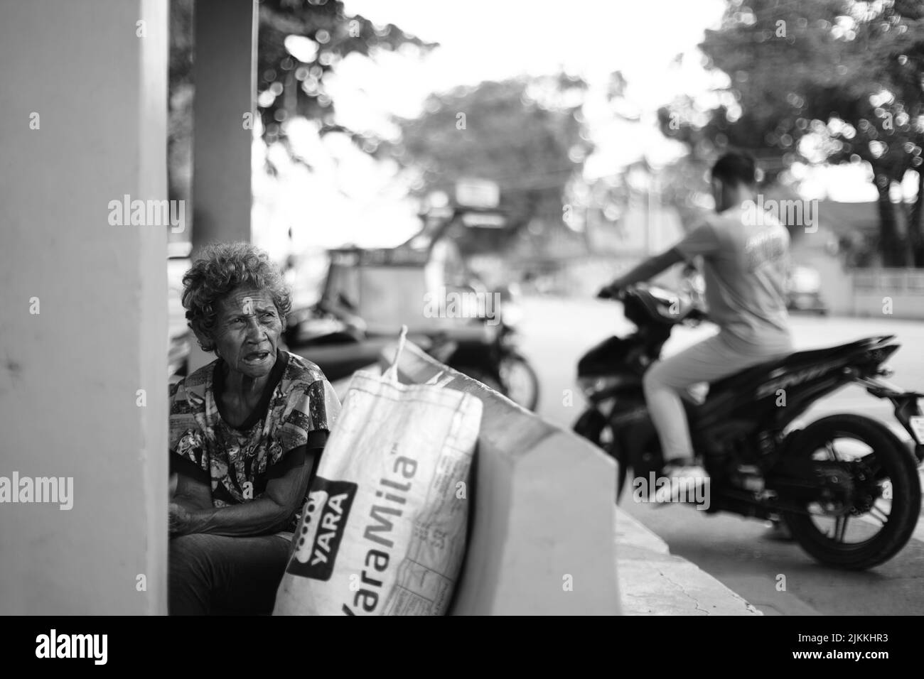 Un colpo in scala di grigi di una donna anziana nelle Filippine con un uomo che guida una motocicletta dietro di lei Foto Stock