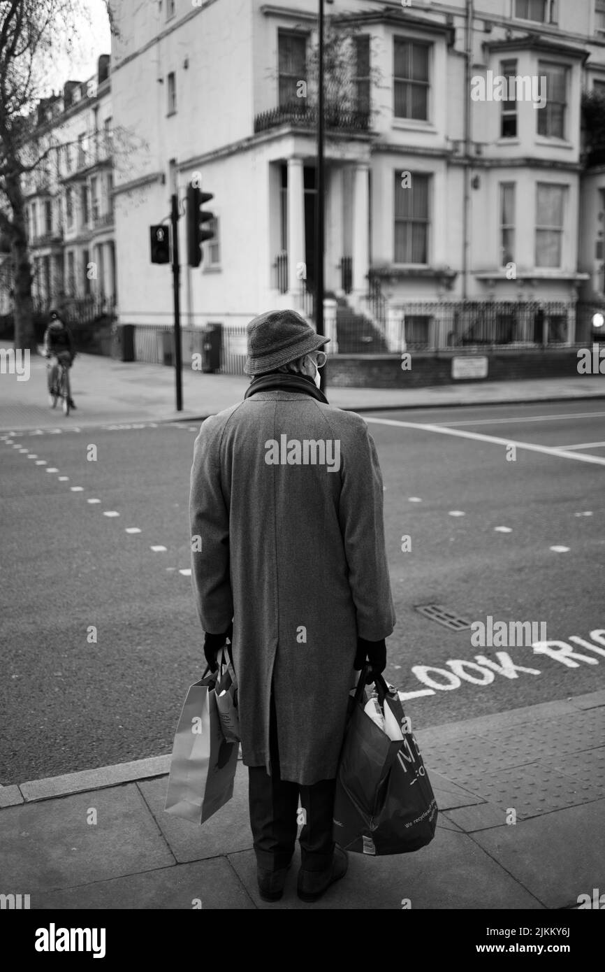Un'immagine in scala di grigi di un vecchio uomo con pacchetti in piedi in una strada su uno sfondo di edifici Foto Stock