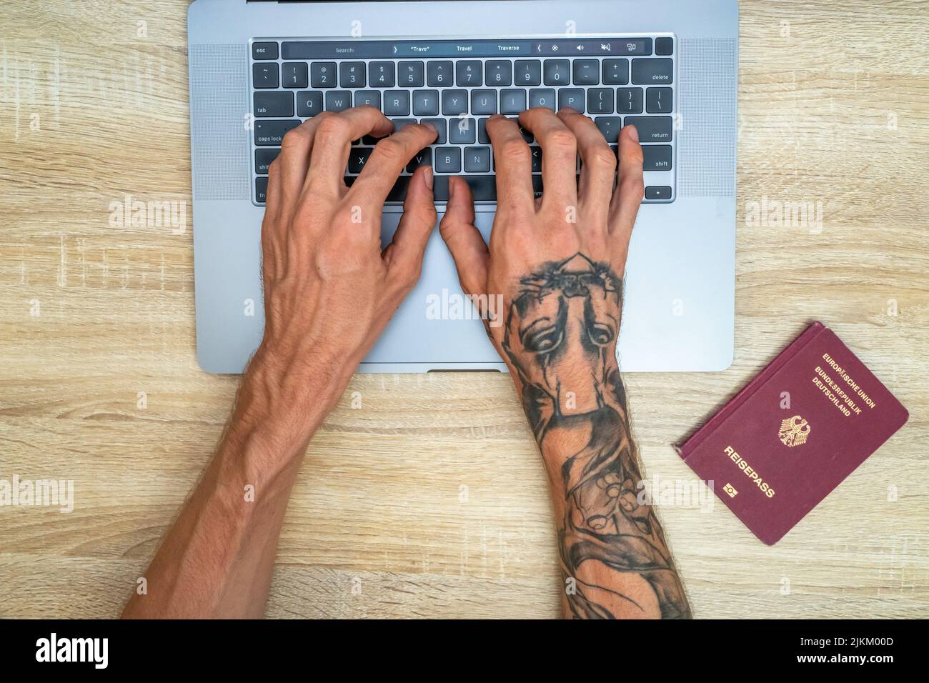 Una vista dall'alto delle mani tatuate che scrivono su un computer portatile in cerca di un volo, con un passaporto tedesco sul lato Foto Stock