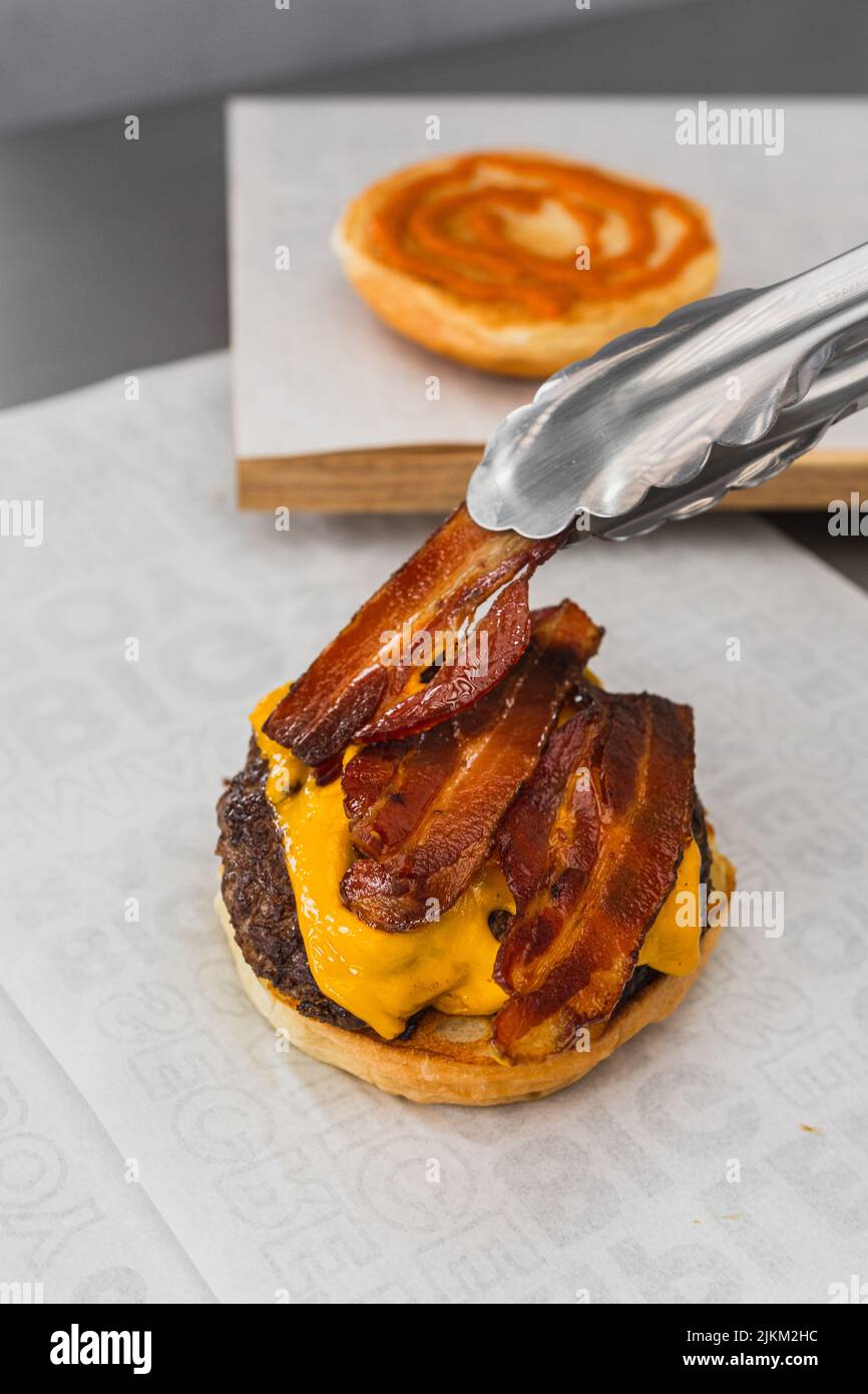 Uno shot verticale di pinze che mette pancetta su un delizioso hamburger succoso Foto Stock