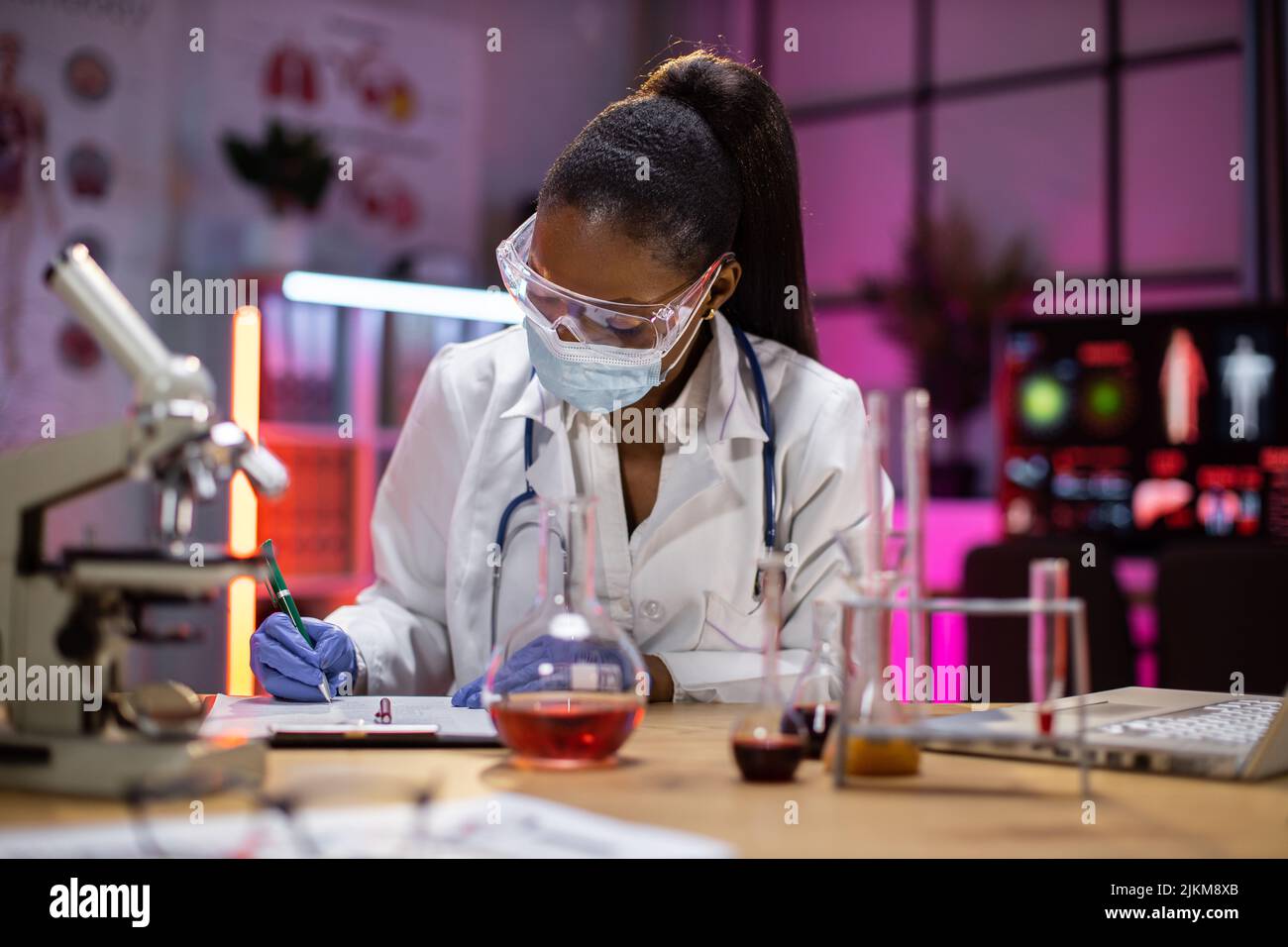 Microbiologo o medico professionista afroamericano usano il riempimento della provetta con sangue e microscopio scrivere risultati di ricerca in laboratorio. Foto Stock
