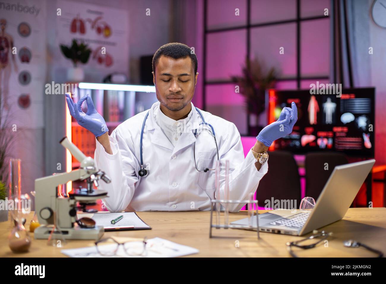 Yong scienziato maschile afroamericano rilassamento pratica yoga indossando camice da laboratorio lavorando in laboratorio mentre si esamina campione di biochimica in provetta e strumenti scientifici. Foto Stock