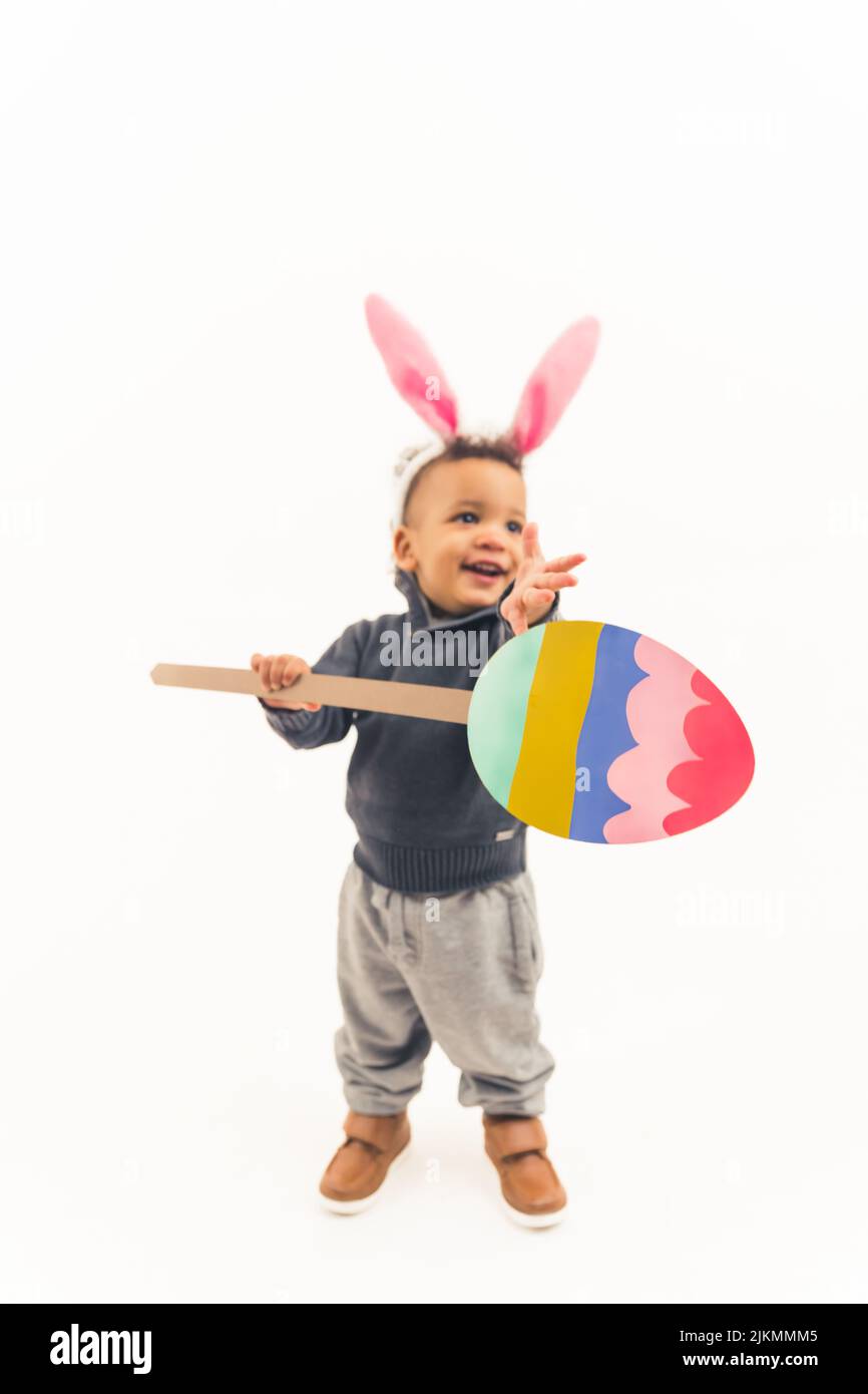 Eccitata carina afroamericana toddler divertirsi con le orecchie di coniglio di pasqua tenendo un grande giocattolo uovo di pasqua sul bastone - isolato. Foto di alta qualità Foto Stock