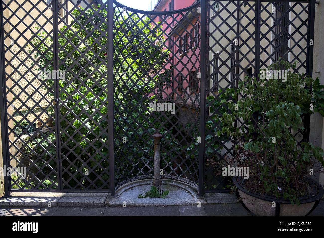 Vecchia fontana pubblica di fronte ad una recinzione con una fila di case e un fico tre su un flusso d'acqua Foto Stock